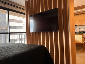 A modern hotel room with a large bed, a flat-screen TV mounted on a wooden panel wall, and a view of the city through a large window. - ID Vida Urbana (Goiânia)