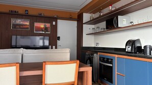A compact kitchen with a microwave, toaster, and coffee maker, a dining area with a sofa and chairs, and a TV. - ID Vida Urbana (Goiânia)