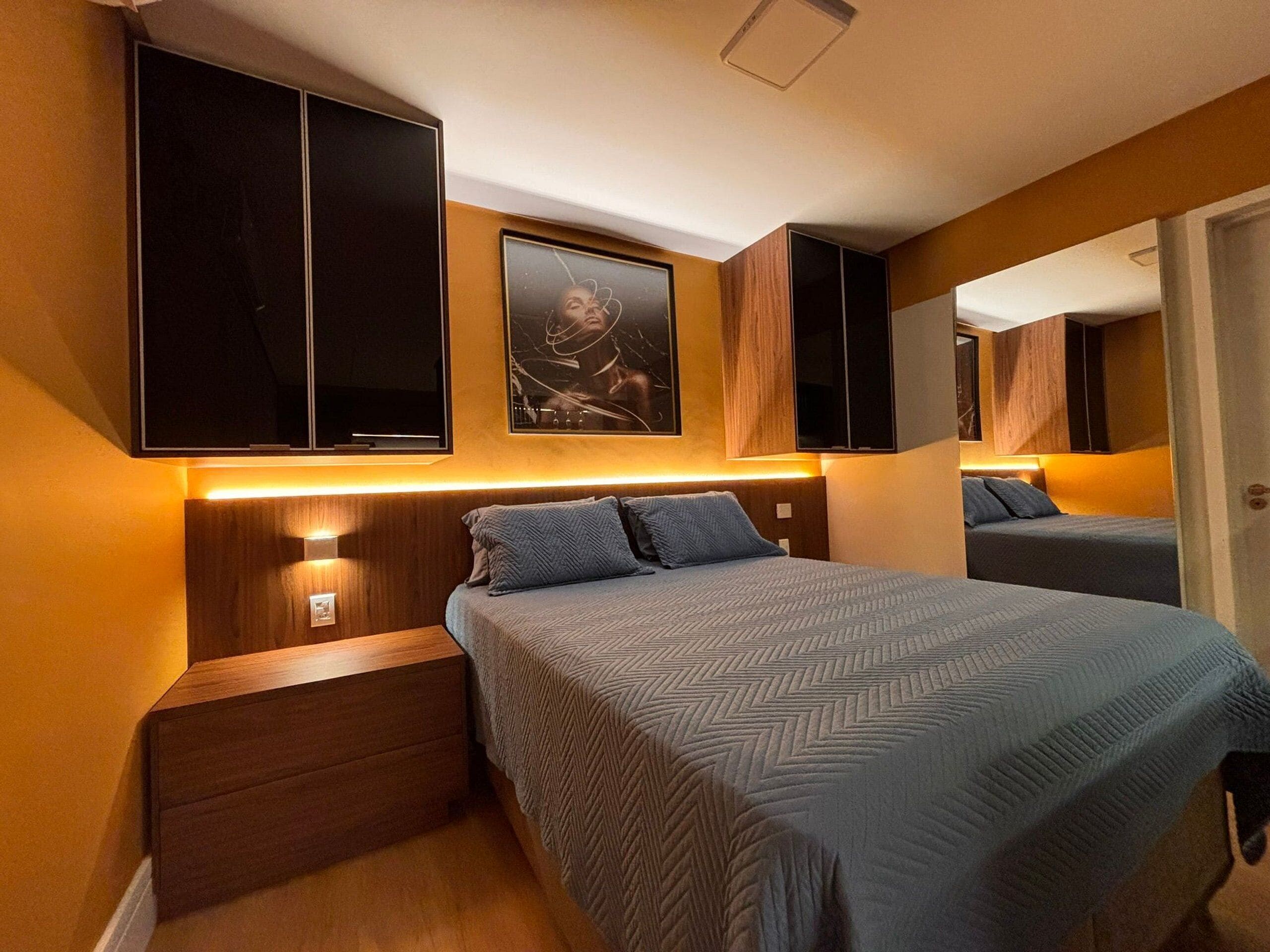 Quarto com cama, mesas de cabeceira, televisão presa à parede, quadro emoldurado e espelho.