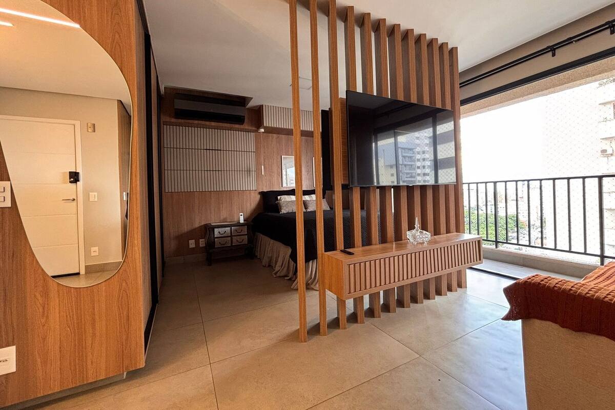 A modern hotel room with a wooden partition, a TV, a balcony with a view, and a small kitchen area.