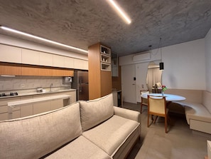 A modern kitchen with a built-in refrigerator, a dining area with a round table and chairs, and a sofa. - ID Vida Urbana (Goiânia)