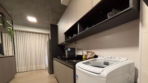 A modern kitchen with a built-in washer, a black countertop, and a window with vertical blinds. - ID Vida Urbana (Goiânia)