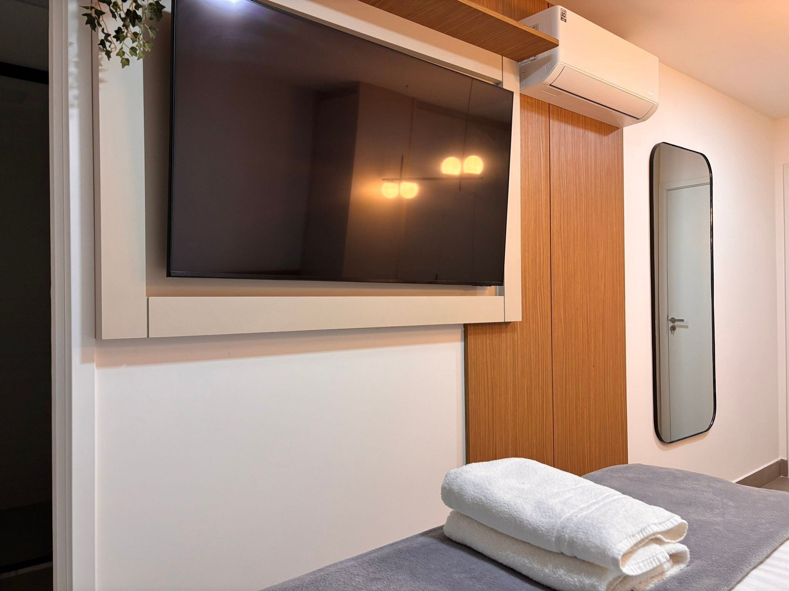 Ein Zimmer mit einem Bett, einem an der Wand befestigten Fernseher, einem Spiegel und einem Handtuch auf dem Bett.