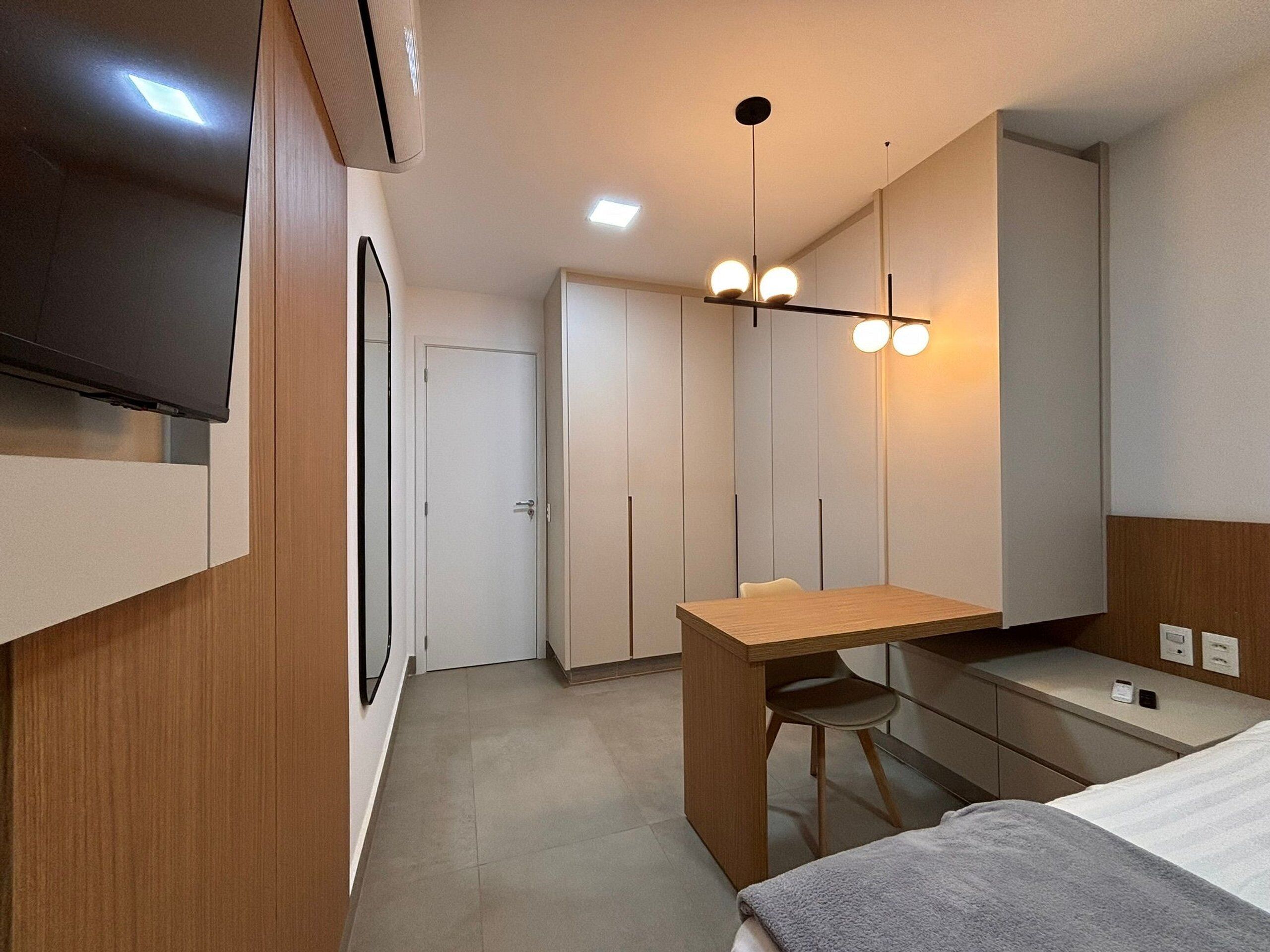 A modern hotel room with a wooden desk, a chair, a bed, a TV, and a ceiling light fixture.