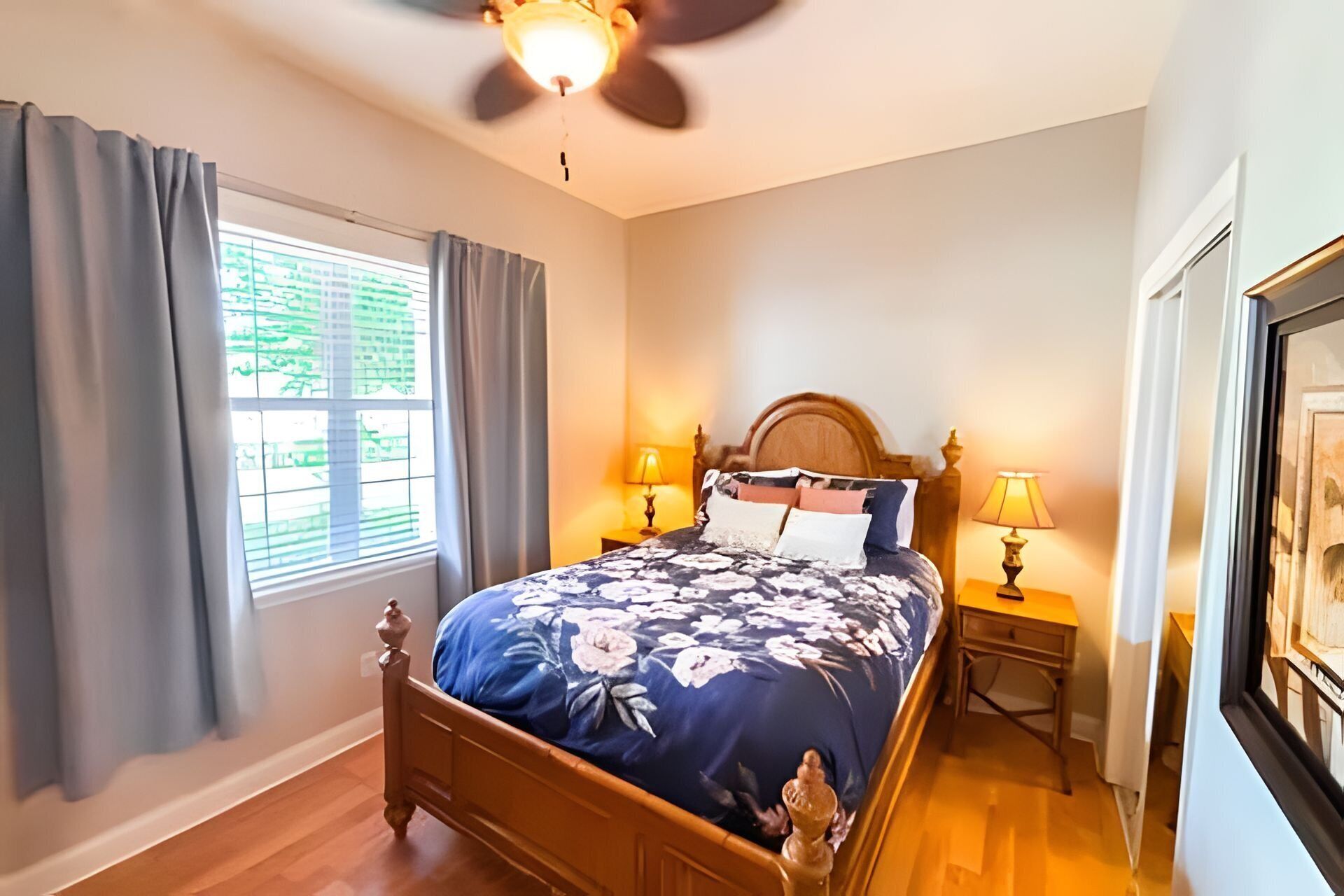 A bedroom with a wooden bed, two bedside tables with lamps, a ceiling fan, and a window with curtains.