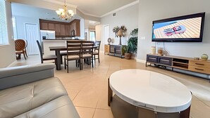 Un salon moderne doté d’un téléviseur à écran plat, d’un coin repas avec une table et des chaises en bois, et d’une cuisine en arrière-plan.