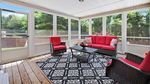 Une terrasse couverte avec un tapis à motifs noir et blanc, des meubles en osier rouge et noir, et une vue sur une clôture en bois et des arbres.