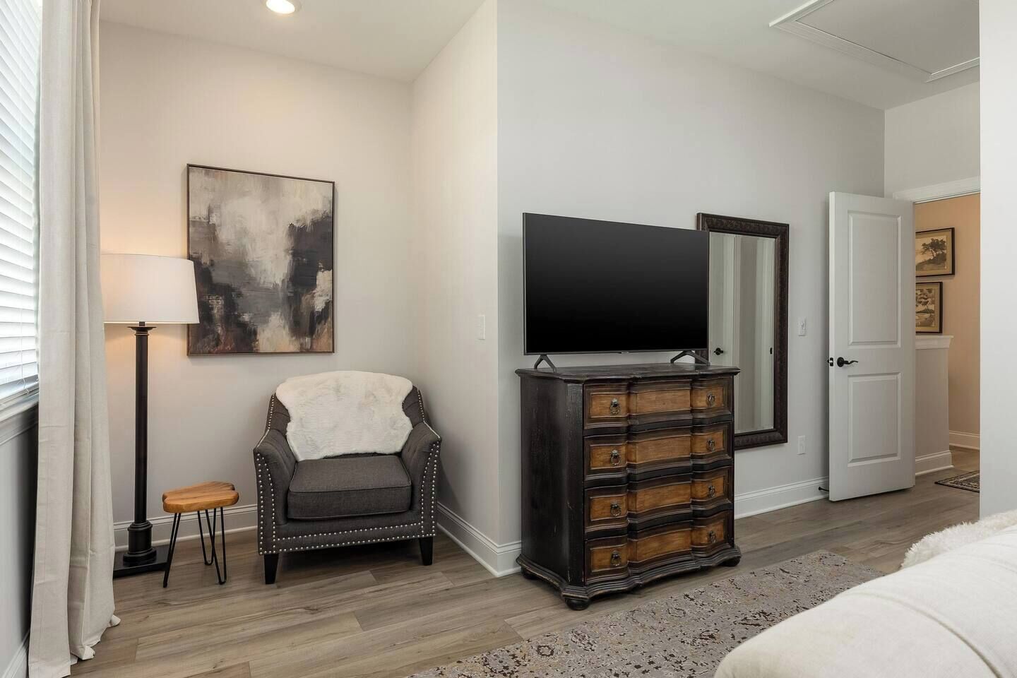 A bedroom with a large flat-screen TV, a dark wooden dresser, a chair with a white fur throw, a lamp, and a door leading to another room.