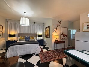A bedroom with a bed, bedside tables, a desk, a chair, a rug, a ceiling light, and a refrigerator.