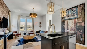 A modern kitchen with a black countertop, stainless steel sink, and a large island. There is a dining area with a round table and chairs, and a living room with a sofa and a large window. - NEW!  Sleek Rooftop Retreat - Sleeps 10 (Nashville)
