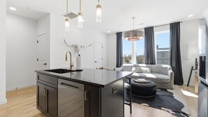 Ett modernt kök med en köksö i mitten, en matplats med soffa och tv samt ett stort fönster med gardiner.