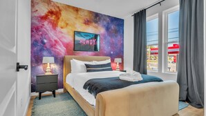 A bedroom with a galaxy-themed wall, a bed with white bedding, two bedside tables with lamps, and a view of a gas station through the window. - Rooftop Views + Relaxing Hot Tub & Fire Pit (Nashville)