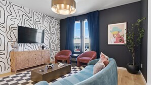Ein modernes Wohnzimmer mit Schachbrettmuster-Boden, einer Couch, zwei Sesseln, einem Fernseher, einem Schrank und einem großen Fenster mit Vorhängen.