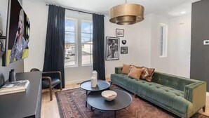 Un salon moderne avec un canapé vert, deux petites tables rondes, une grande fenêtre avec des rideaux et un téléviseur fixé au mur.