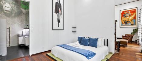 Habitación de hotel moderna con una cama grande, un baño con ducha de vidrio y un área de comedor con mesa y sillas.