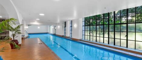 An indoor swimming pool with clear glass walls, a tiled floor, and potted plants.