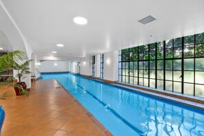 An indoor swimming pool with clear glass walls, a tiled floor, and potted plants.