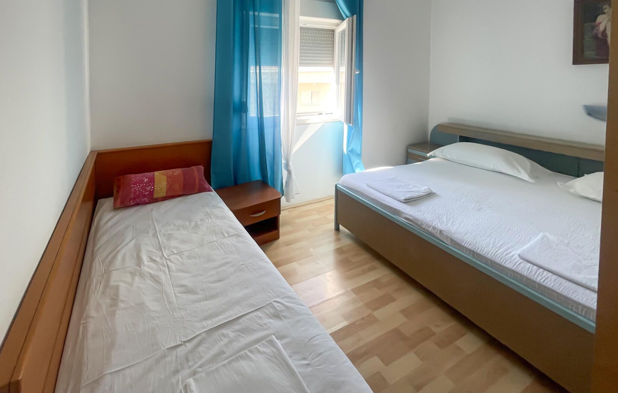 Une chambre avec deux lits, un banc en bois, une table de chevet et une fenêtre avec des rideaux bleus.