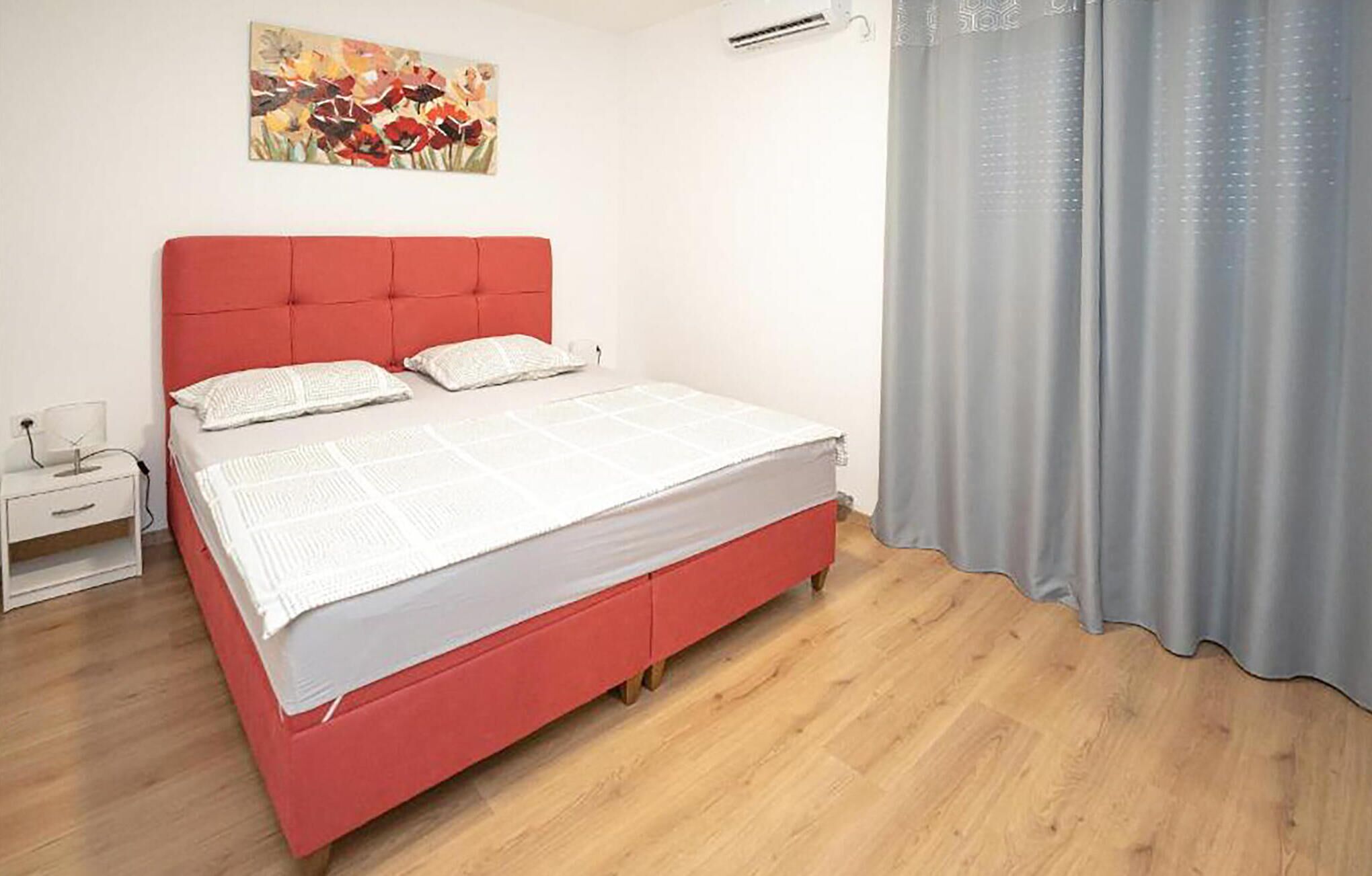 A bedroom with a red bed, white bedding, a nightstand, a painting on the wall, and a window with curtains.