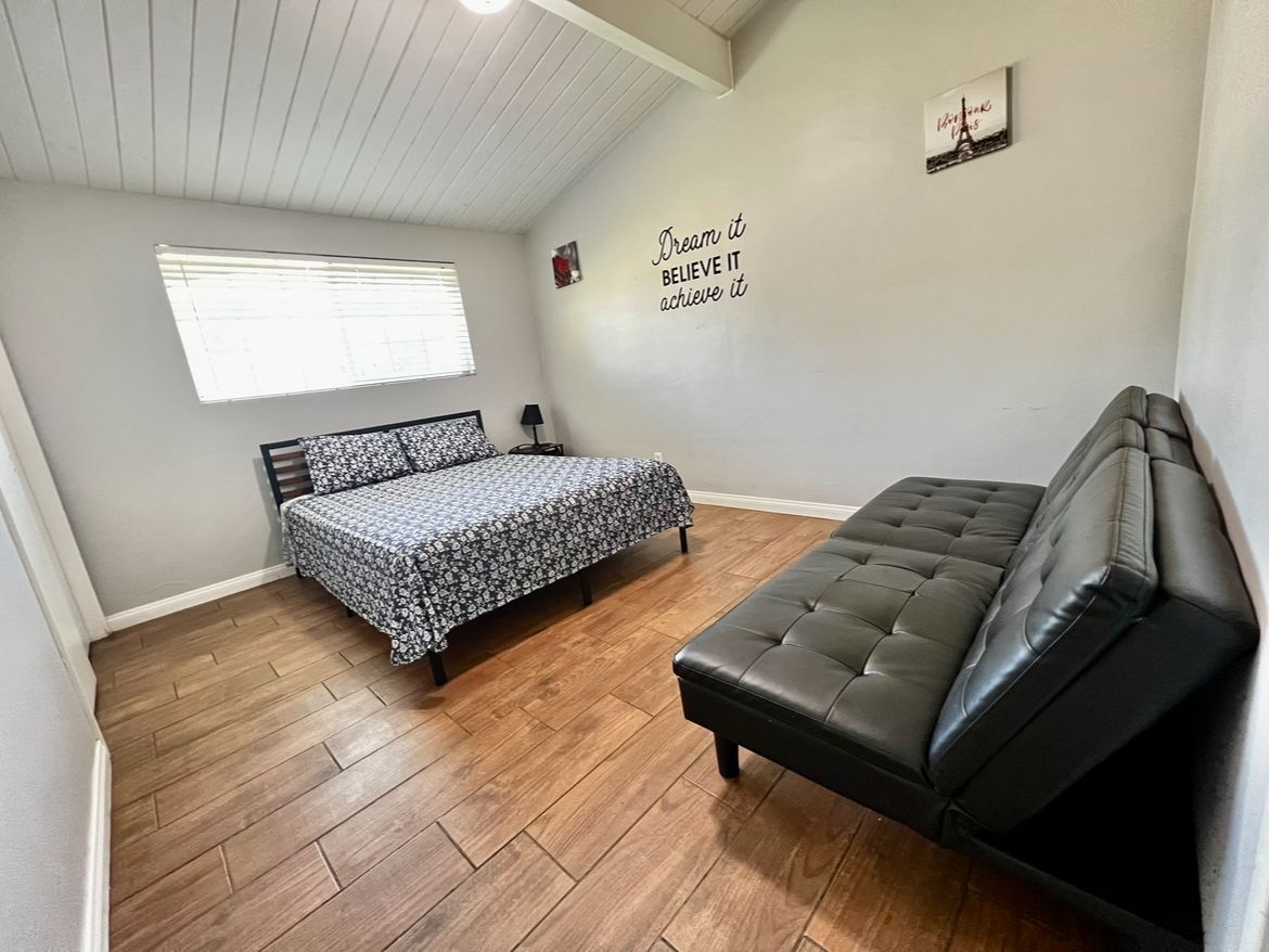 A bedroom with a bed, a sofa, wooden flooring, a window with blinds, and a motivational wall quote.