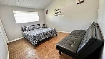 A bedroom with a bed, a sofa, wooden flooring, a window with blinds, and a motivational wall quote.