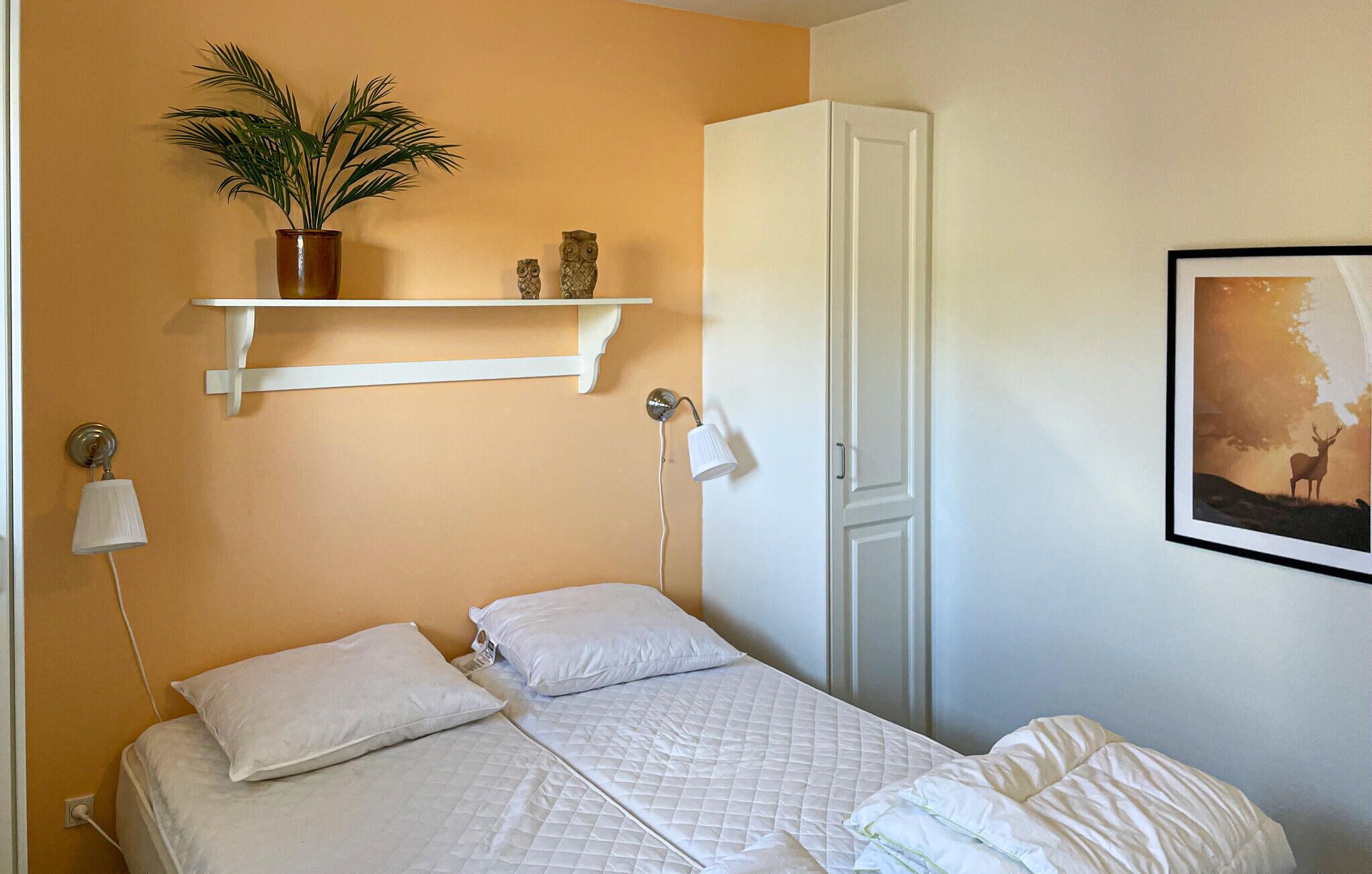 A bedroom with two beds, a shelf with decorative items, a wall-mounted lamp, and a framed picture.
