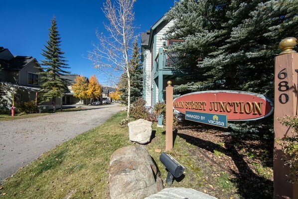 A street sign for Main Street Junction managed by Vacasa, with a clear road, residential houses, and a large evergreen tree.
