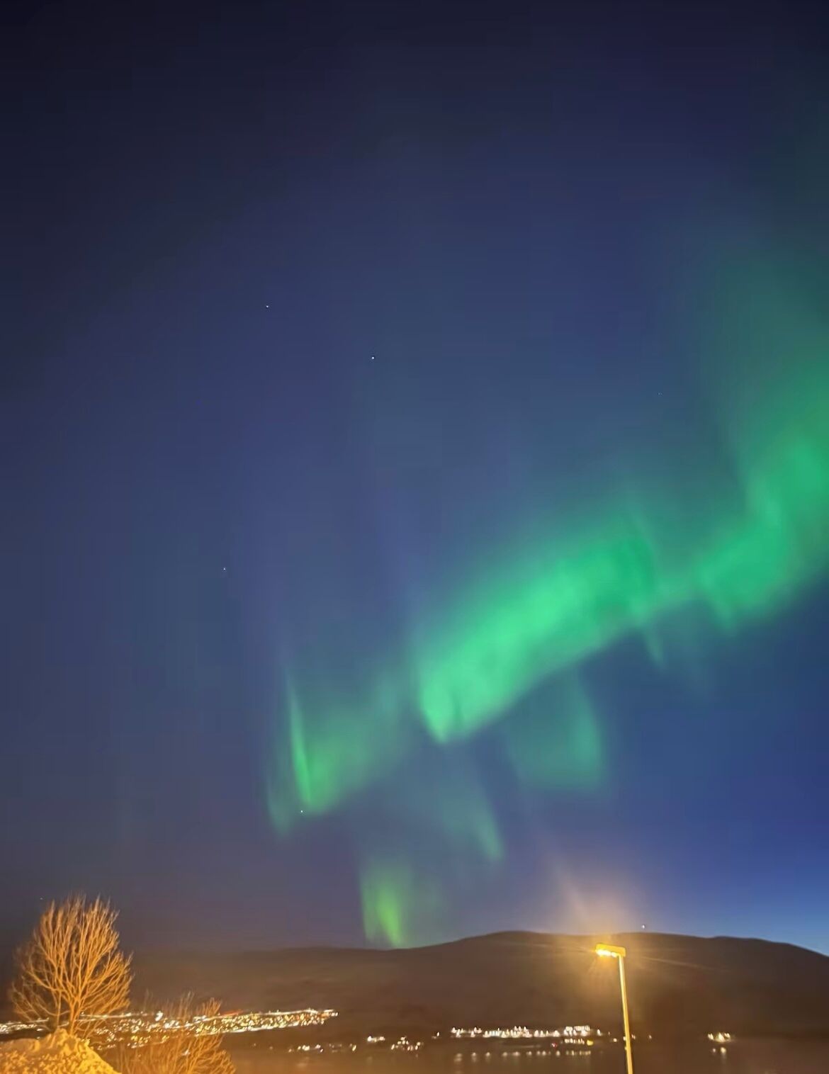 A night view of the Northern Lights over a town with visible buildings and a hill.
