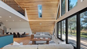 A spacious living area with a wooden ceiling, a grey sofa, a coffee table, and a view of a road through large windows. - Bend Retreat - Hot Tub, Fire Pit & Waterfront (Bend)
