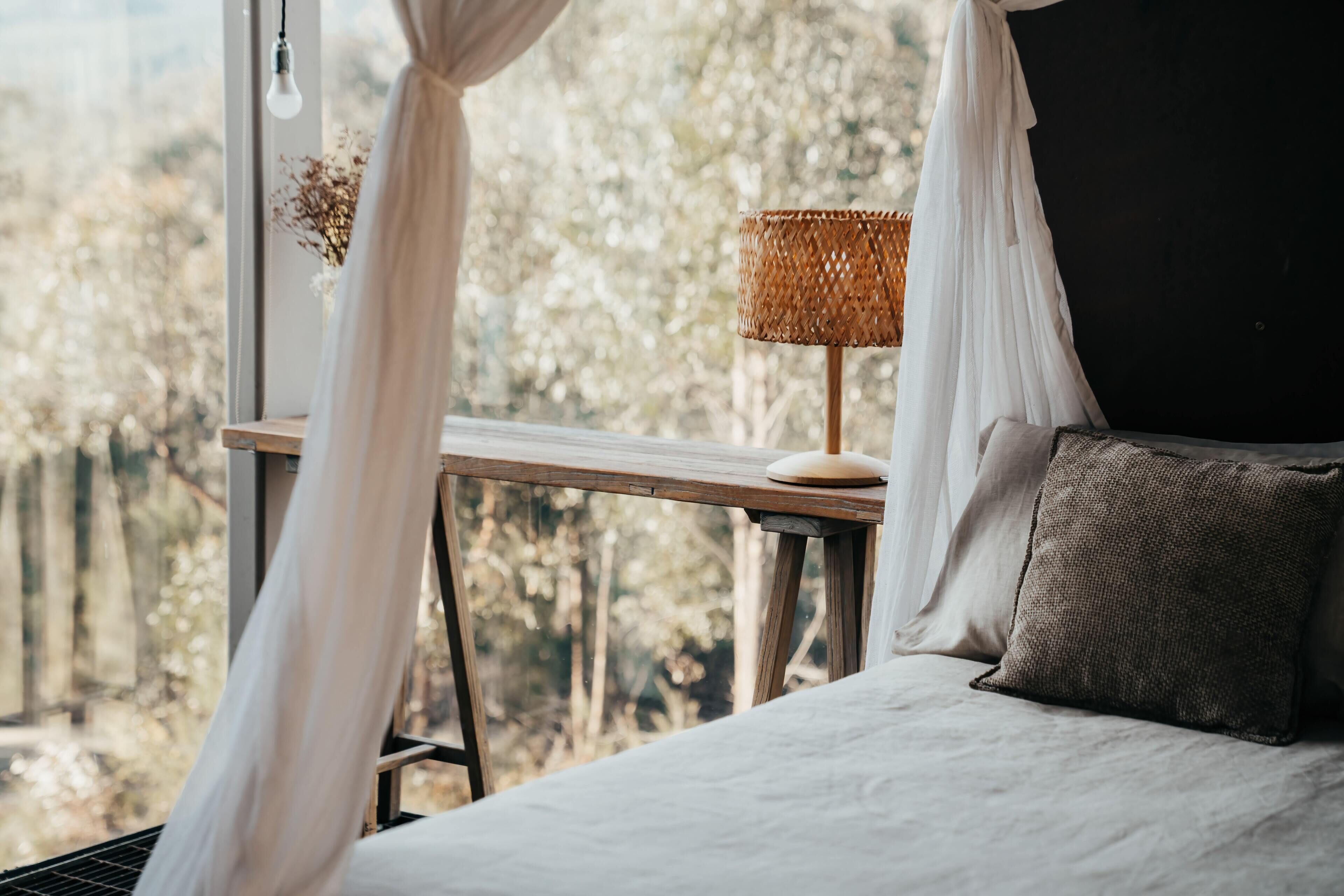 A bedroom with a bed, a bedside table with a lamp, and a view of trees through a window.