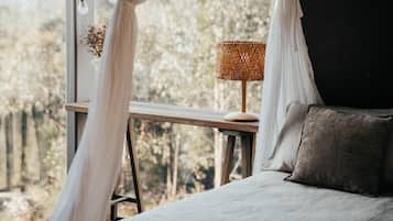 A bedroom with a bed, a bedside table with a lamp, and a view of trees through a window.