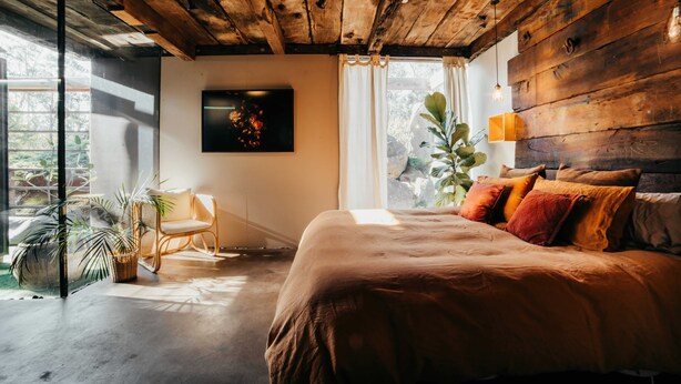 Une chambre avec un plafond en bois, un lit recouvert de plusieurs coussins, une chaise et une grande fenêtre avec des rideaux.