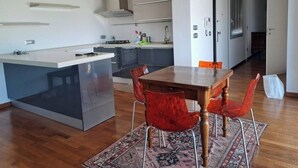 Moderni keittiö, jossa on keskeinen saareke, ruokailutila puupöydällä ja punaisilla tuoleilla sekä käytävä, joka johtaa makuuhuoneeseen.