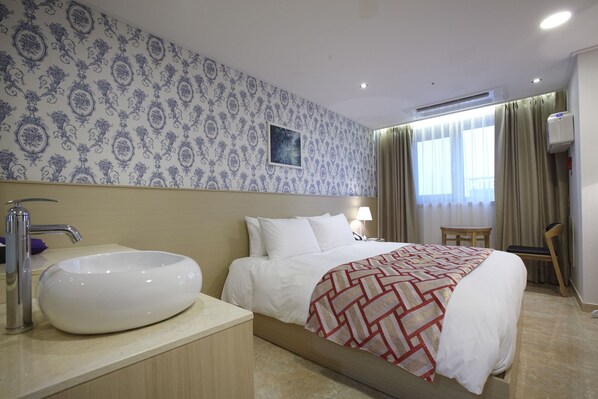 A hotel room with a large bed, a bedside table with a white basin, a chair, a small table, and a window with curtains.