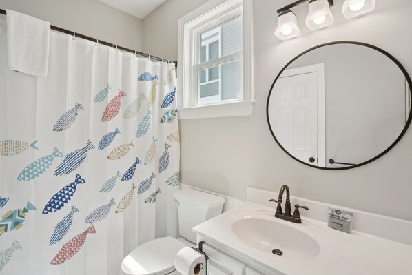 Un baño con cortina de ducha con motivos de peces, un inodoro, un lavabo con grifo, un espejo y una ventana sobre el lavabo.