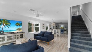 A spacious living room with a large TV, a dark blue sofa, and a staircase leading to the upper floor. - Stylish 3-Story Home w/ Heated Pool & Palm Views (Naples)