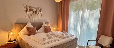A bedroom with a bed, bedside tables, a chair, a hanging light fixture, and a window with curtains.