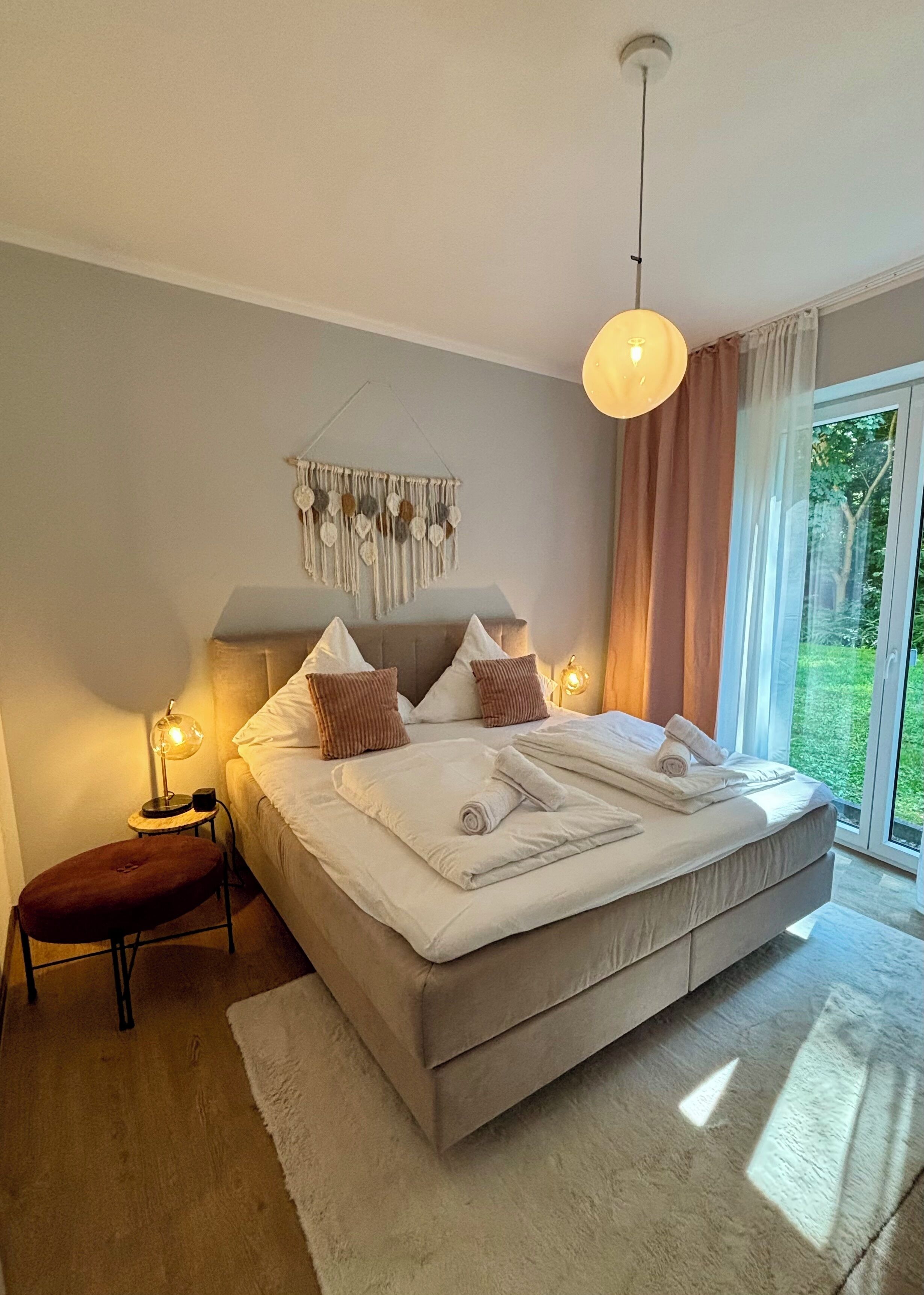 Un dormitorio moderno con una cama grande, lámparas de noche, un adorno de macramé en la pared y vistas a la vegetación por la ventana.
