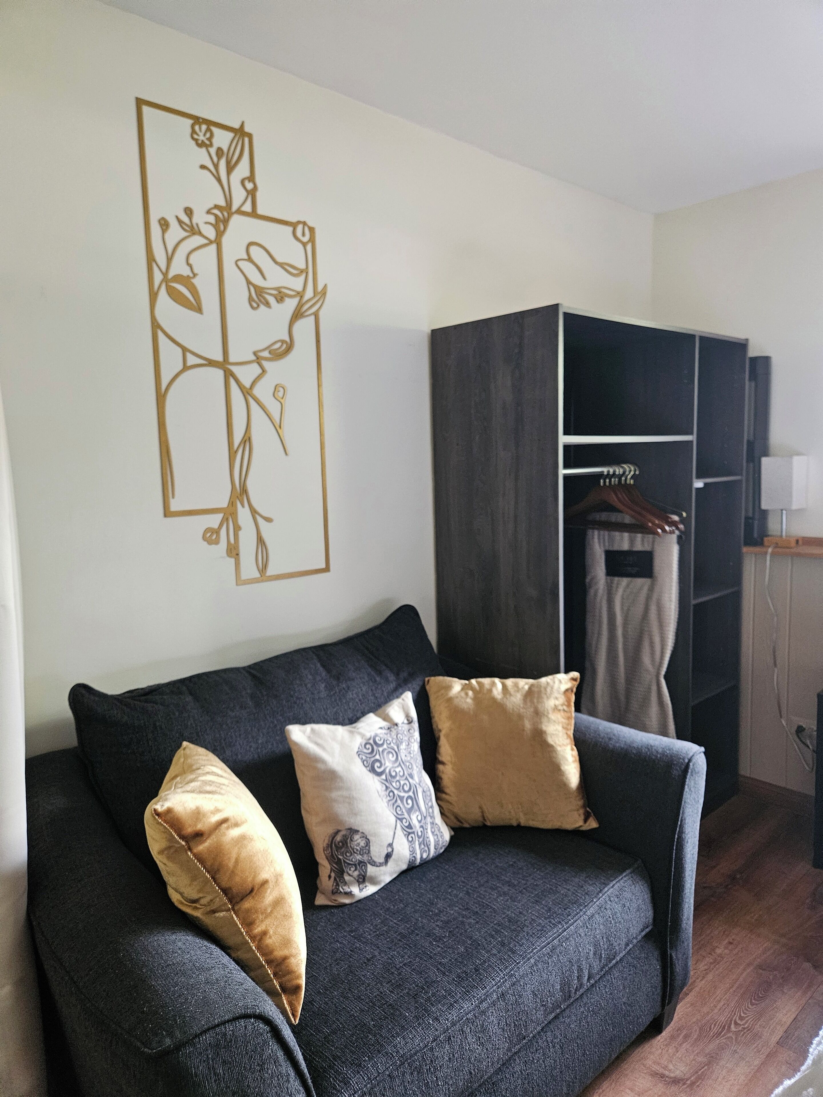 A dark grey sofa with three cushions, a wall art piece, and a wardrobe in a room with wooden flooring.