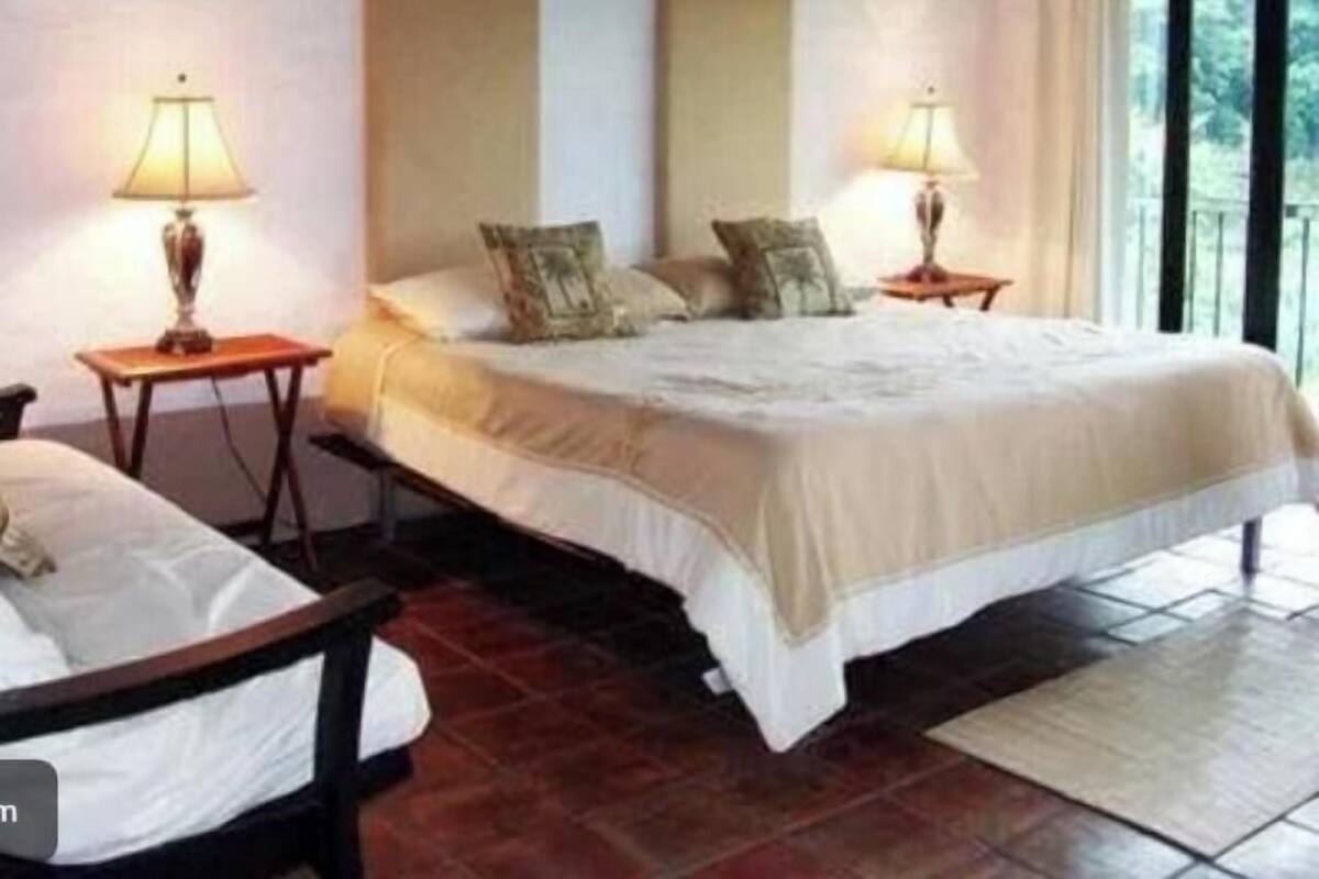 Habitación de huéspedes con una cama grande, dos mesitas de noche con lámparas, una silla y vistas a la vegetación por la ventana.