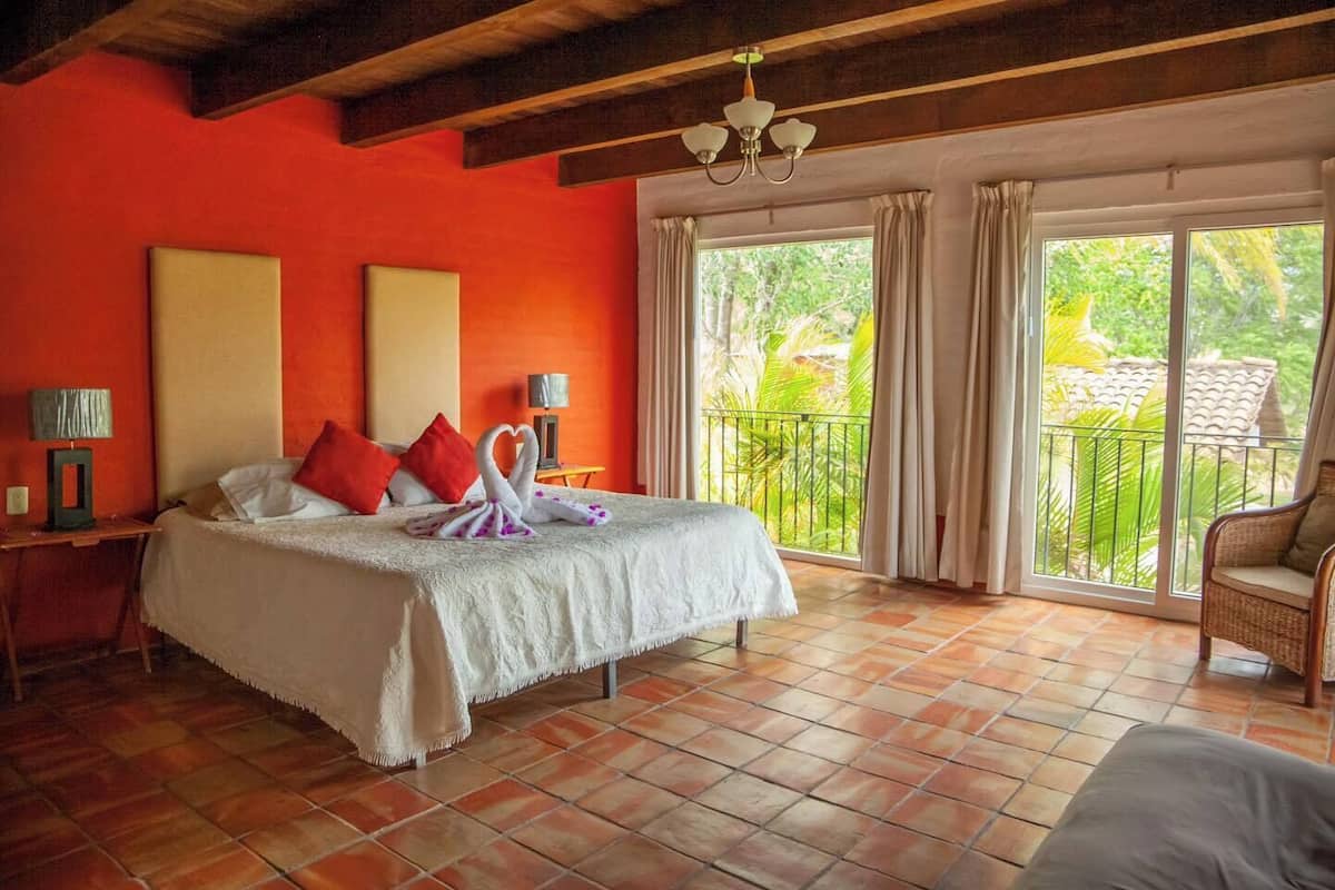 Habitación de hotel con una cama grande, paredes rojas, un balcón con cortinas y vistas a zonas verdes.