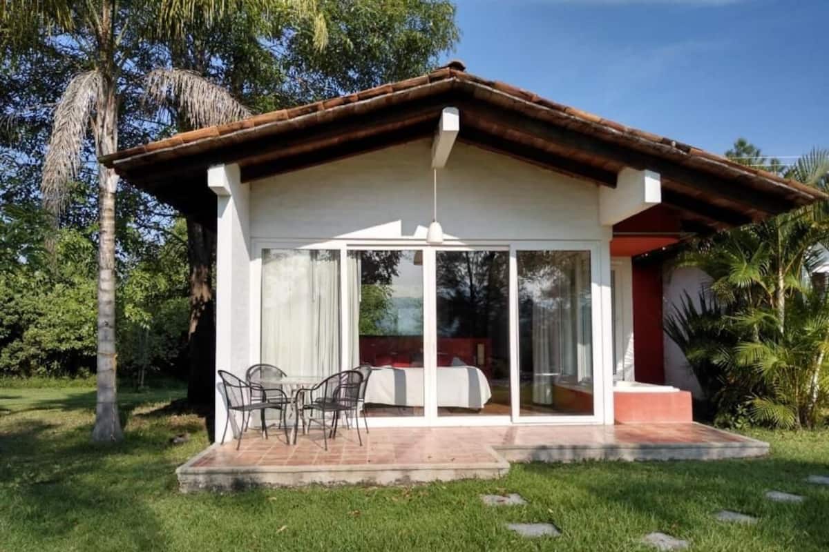 Una casa de una planta con patio, un comedor con sillas y una cama visible a través de las puertas de cristal.