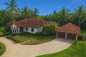A two-story building with a red-tiled roof, surrounded by lush greenery and palm trees.