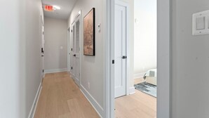 Un couloir doté d’un plancher en bois, de murs blancs et d’une porte donnant sur une chambre à coucher où se trouvent un lit et un tapis.