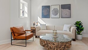 Un salon moderne avec un fauteuil en cuir, une table basse ronde en verre, un canapé beige et trois œuvres d’art encadrées accrochées au mur.