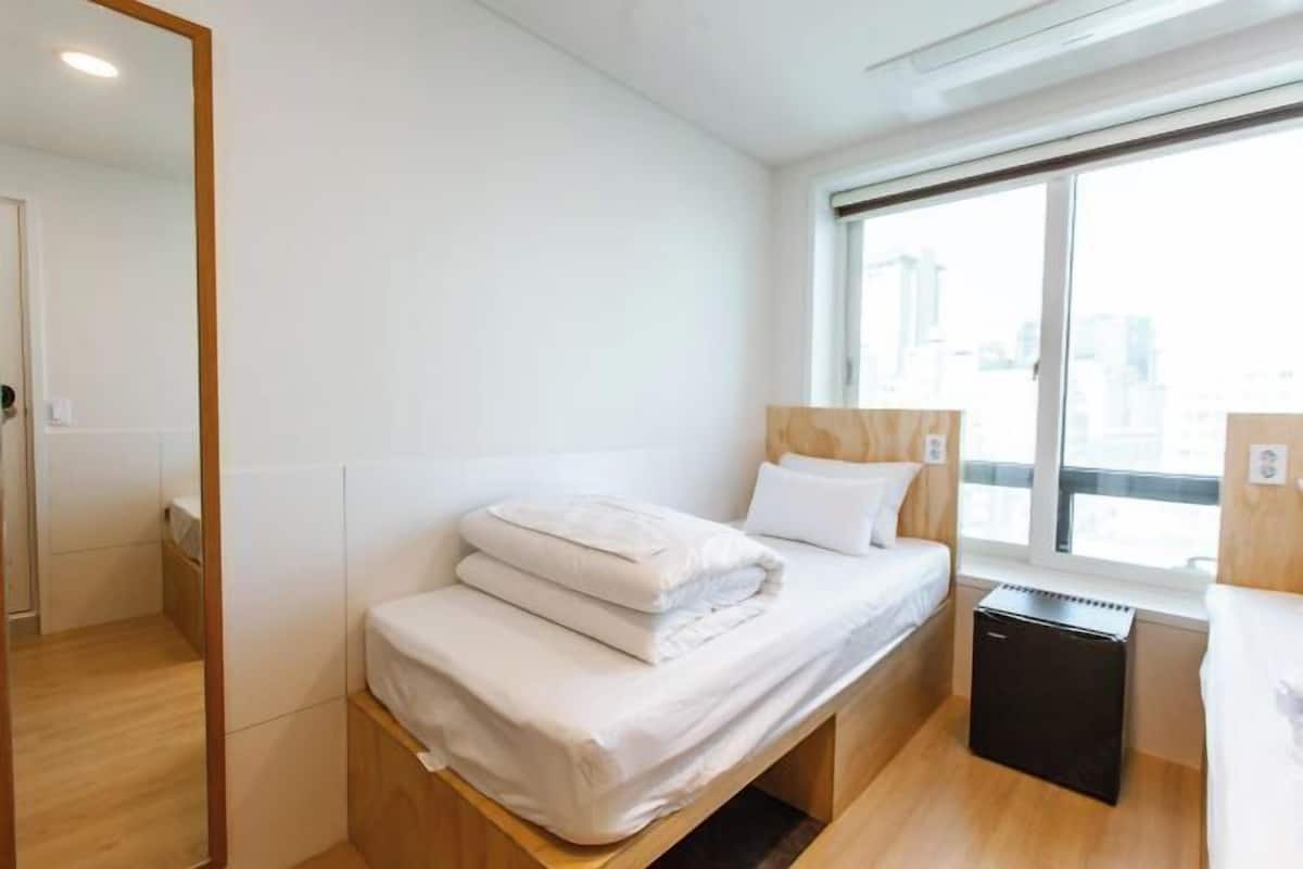 Habitació d'hotel senzilla amb un llit individual, un escriptori petit i una finestra que ofereix vistes a la ciutat.