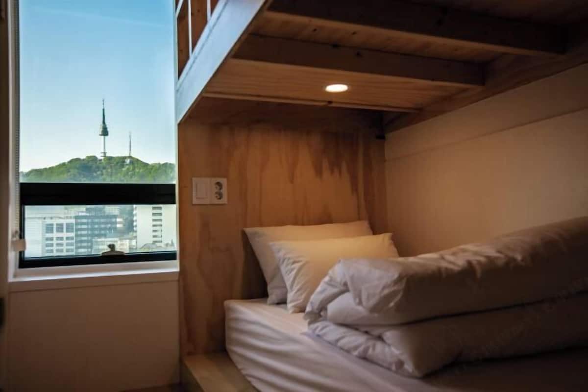 Habitació petita i acollidora amb un llit individual, una finestra que mostra una vista urbana i panells de fusta.