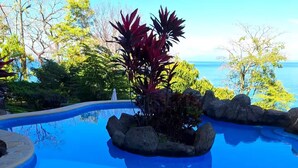 Una piscina azul cristalina con una formación rocosa y plantas, rodeada de árboles y vistas al océano.