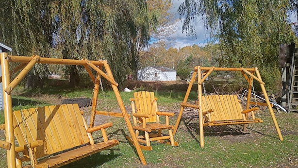 Een houten schommel met twee schommelstoelen en twee stoelen in een grasveld met bomen en een huis op de achtergrond.