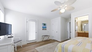 Un dormitorio con cama, escritorio, silla, ventilador de techo y baño con espejo.